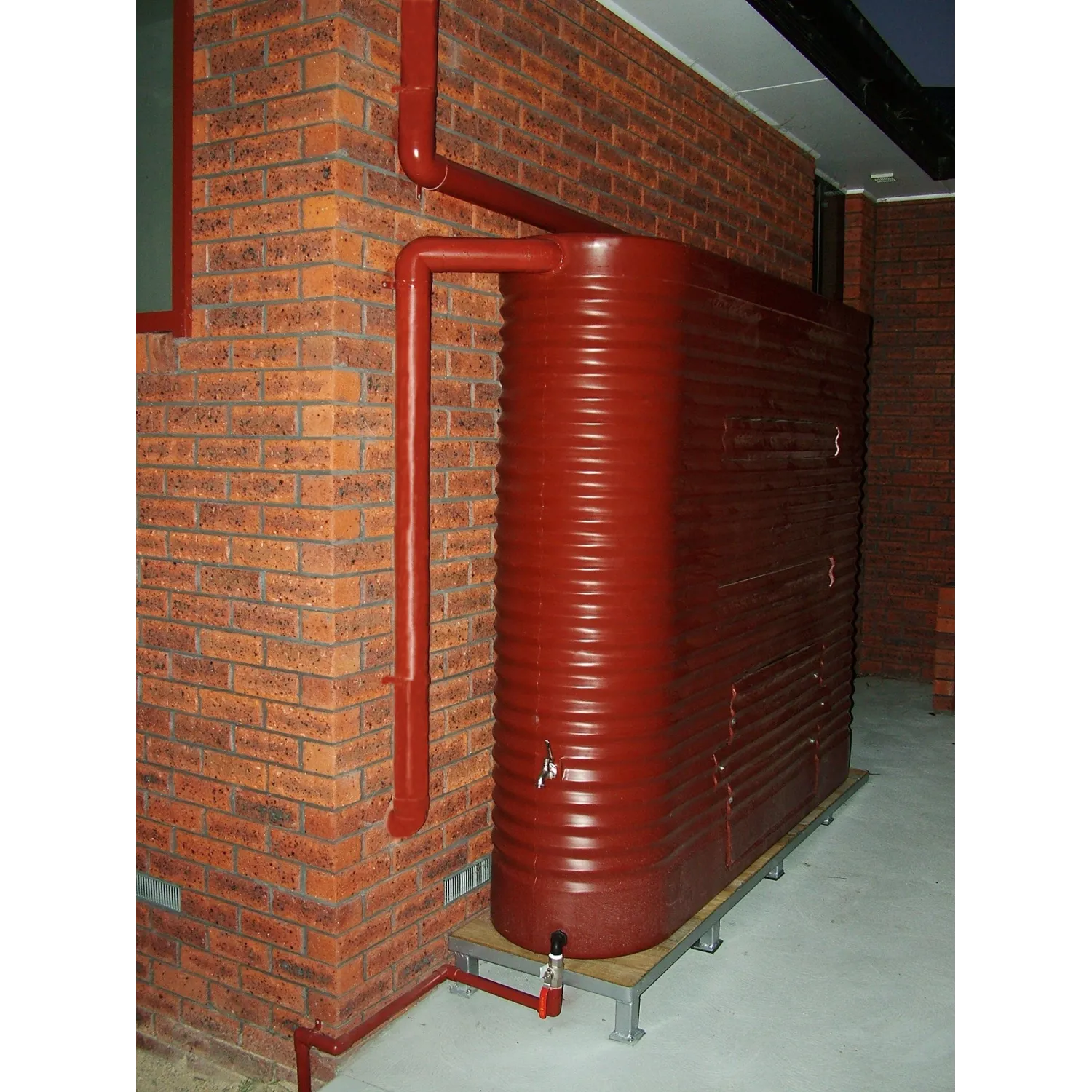 2000 Ltr Corrugated Slimline Tank on a modern metal and timber base raised over concrete next to brown bricks.